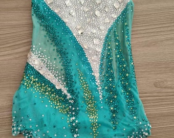 May include: A turquoise and white leotard with sparkling embellishments. The sleeveless garment features a V-neck design with a mesh overlay and is adorned with rhinestones and sequins in a decorative pattern. The leotard is designed for gymnastics or dance.