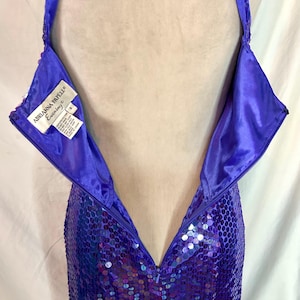 May include: A close-up of a purple sequined evening gown with a halter neckline. The dress has a zipper down the back and a label that reads "Adrianna Papell Evening". The sequins create a shimmering effect.
