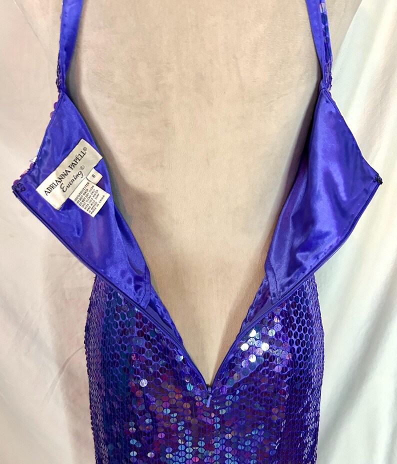 May include: A close-up of a purple sequined evening gown with a halter neckline. The dress has a zipper down the back and a label that reads "Adrianna Papell Evening". The sequins create a shimmering effect.