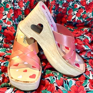 May include: A pair of platform sandals with a wooden wedge heel. The sandals have a light pink, criss-cross strap design and a clear sole decorated with red and white hearts. The wooden wedge has a heart-shaped cutout. The sandals are on a floral background.