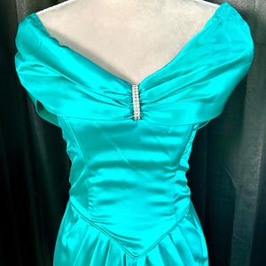 May include: A teal-colored formal dress with an off-the-shoulder neckline. The dress features a fitted bodice and a flowing skirt. A sparkling rhinestone embellishment adorns the center of the neckline. The dress is made of a shiny, satin-like fabric.