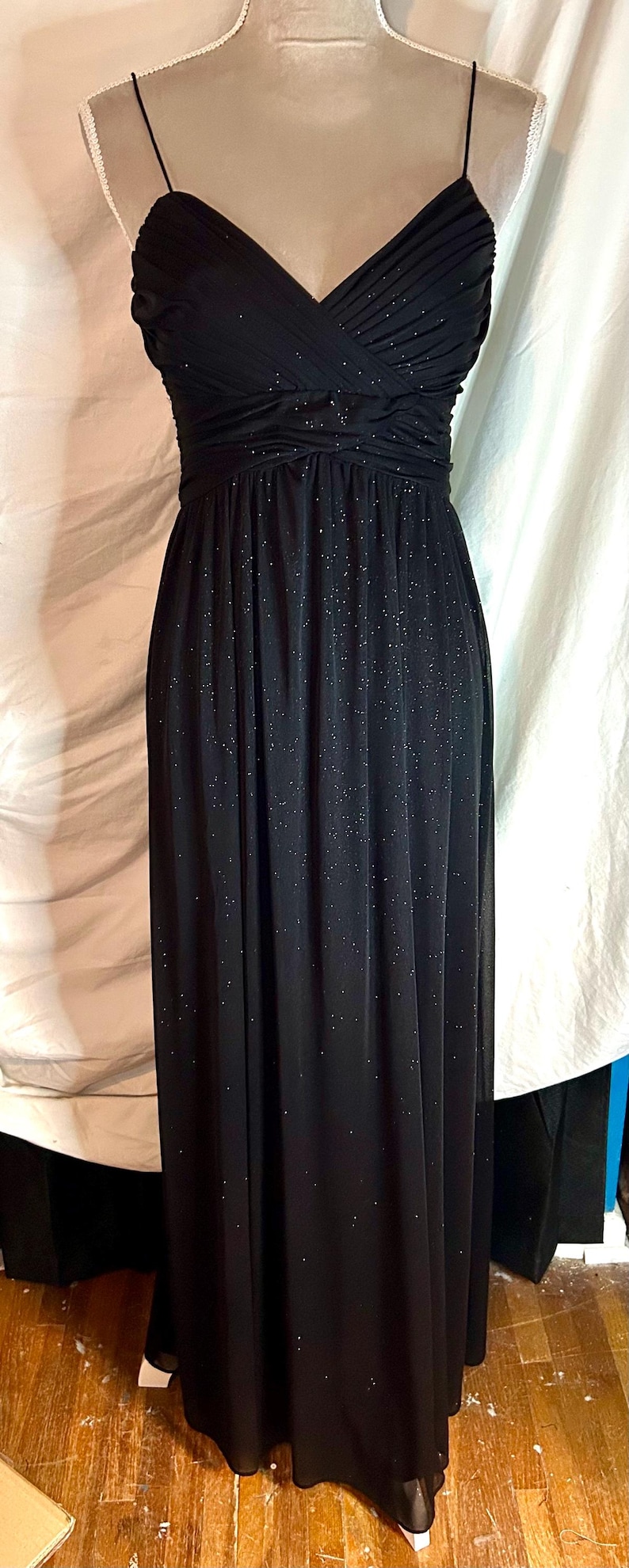 Puede incluir: Vestido de noche negro largo con tejido brillante y con purpurina. El vestido presenta un escote en V, tirantes finos y un corpi&ntilde;o fruncido. La falda llega hasta el suelo.