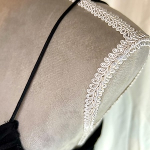 Puede incluir: Primer plano de un vestido de terciopelo negro sobre un maniqu&iacute;. El vestido tiene tirantes finos y un escote pronunciado. El hombro est&aacute; adornado con encaje blanco. El maniqu&iacute; es de color beige neutro.