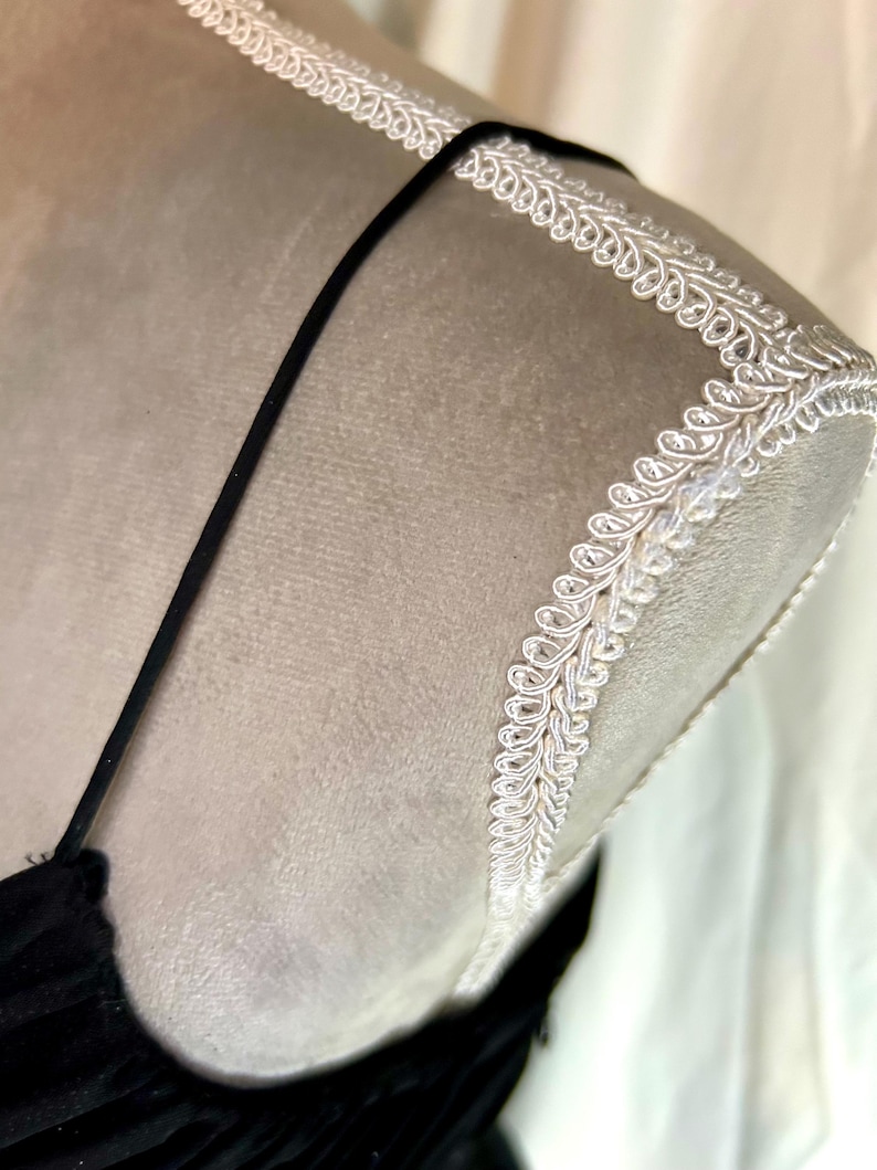 Puede incluir: Primer plano de un vestido de terciopelo negro sobre un maniqu&iacute;. El vestido tiene tirantes finos y un escote pronunciado. El hombro est&aacute; adornado con encaje blanco. El maniqu&iacute; es de color beige neutro.
