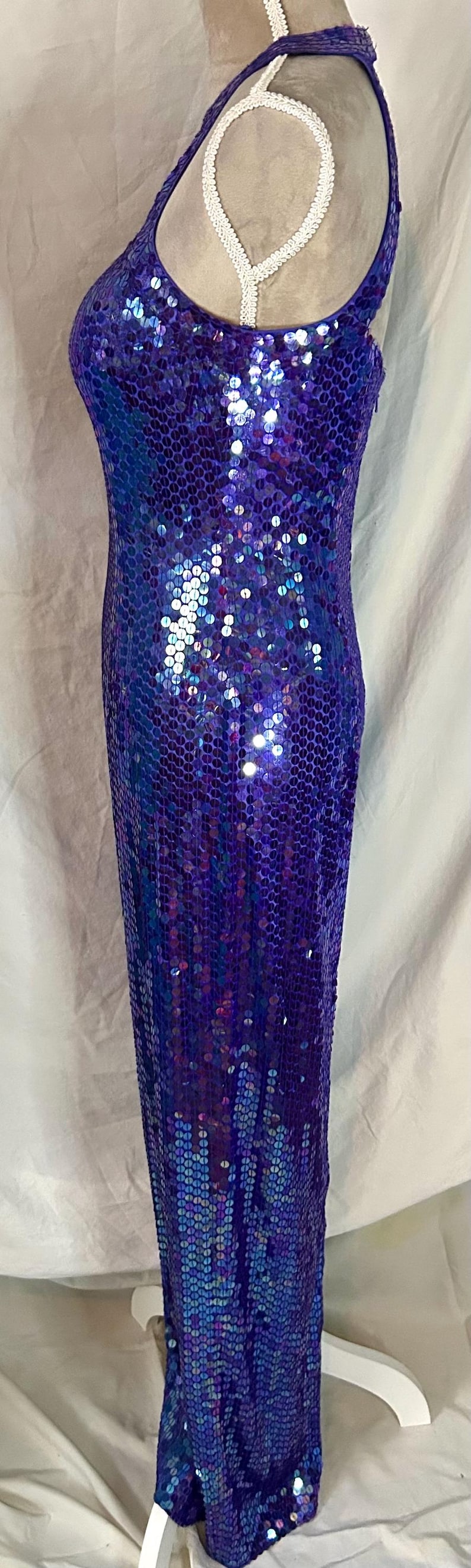 May include: A floor-length, sleeveless gown covered in iridescent blue sequins. The dress has a halter neckline and a form-fitting silhouette. The sequins create a shimmering effect, reflecting light. The dress is displayed on a mannequin.