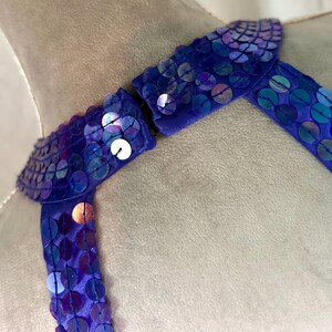 May include: Close-up of a blue sequined accessory. The accessory is made of a blue fabric and is covered in iridescent sequins. The sequins reflect shades of purple, blue, and green. The accessory appears to be a collar or neckline.