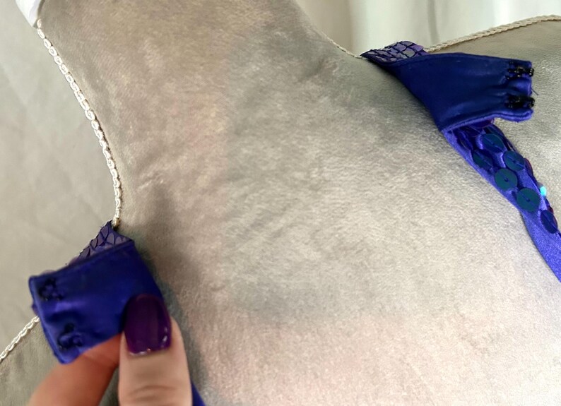 May include: Close-up of a blue garment with sequin and beaded details. The garment has a soft, velvety texture and is adorned with small, round sequins. The straps are embellished with sequins and beads.