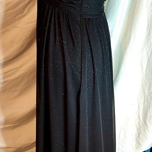 Puede incluir: Un vestido de noche negro con una capa brillante y brillante. El vestido presenta un corpi&ntilde;o fruncido, una falda fluida y delicados tirantes finos. El vestido es largo y tiene cierre de cremallera.