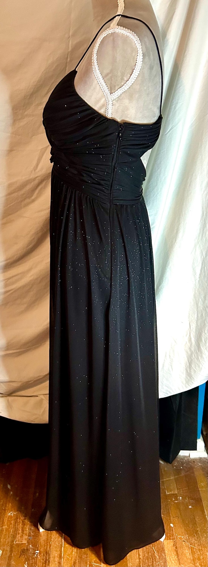 Puede incluir: Un vestido de noche negro con una capa brillante y brillante. El vestido presenta un corpi&ntilde;o fruncido, una falda fluida y delicados tirantes finos. El vestido es largo y tiene cierre de cremallera.