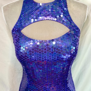 May include: A sleeveless, halter-style dress covered in iridescent blue sequins. The dress features a keyhole cutout at the chest and a form-fitting silhouette. The sequins reflect light, creating a shimmering effect.