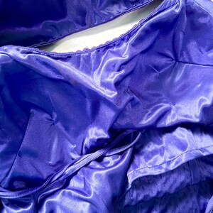 May include: Close-up of a vibrant purple satin garment, possibly a dress or top, with a shiny, flowing texture. The fabric is gathered and draped, with visible seams and a tie detail. The overall look suggests formal wear.
