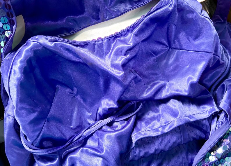 May include: Close-up of a vibrant purple satin garment, possibly a dress or top, with a shiny, flowing texture. The fabric is gathered and draped, with visible seams and a tie detail. The overall look suggests formal wear.
