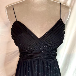 Puede incluir: Un vestido de noche negro con escote en V y tirantes finos. El corpi&ntilde;o est&aacute; fruncido y drapeado, con cintura entallada y falda fluida. La tela tiene un brillo sutil.