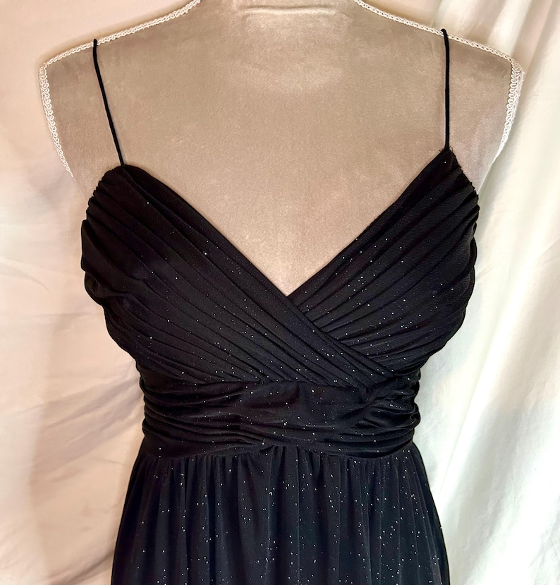 Puede incluir: Un vestido de noche negro con escote en V y tirantes finos. El corpi&ntilde;o est&aacute; fruncido y drapeado, con cintura entallada y falda fluida. La tela tiene un brillo sutil.