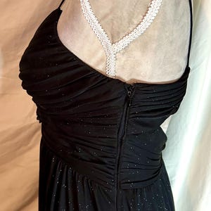 Puede incluir: Vestido de noche negro con corpi&ntilde;o ajustado y falda fluida. El vestido tiene un escote coraz&oacute;n y tirantes finos. La tela est&aacute; adornada con diminutas chispas plateadas. Un detalle de encaje blanco es visible en el hombro.