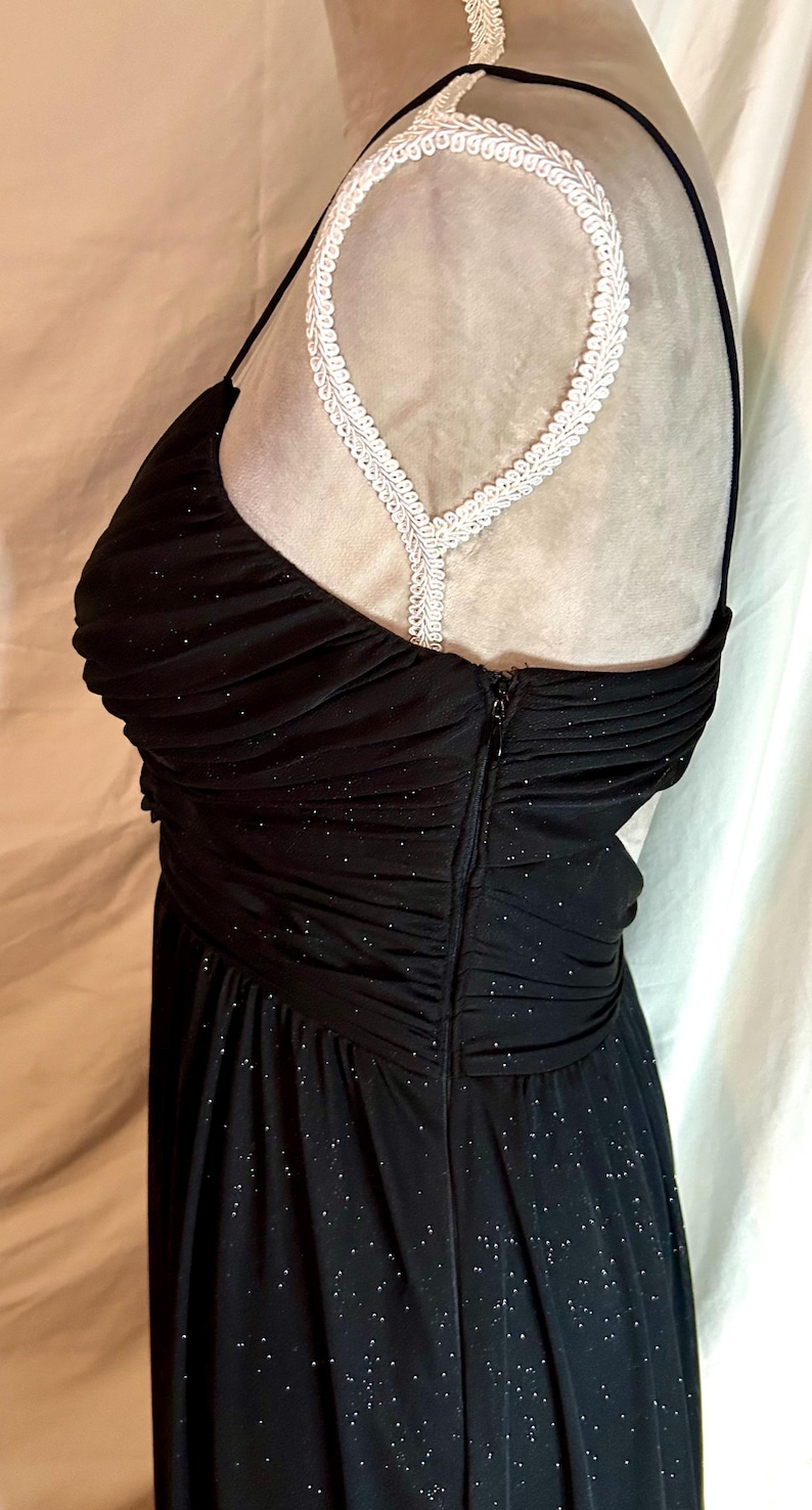 Puede incluir: Vestido de noche negro con corpi&ntilde;o ajustado y falda fluida. El vestido tiene un escote coraz&oacute;n y tirantes finos. La tela est&aacute; adornada con diminutas chispas plateadas. Un detalle de encaje blanco es visible en el hombro.