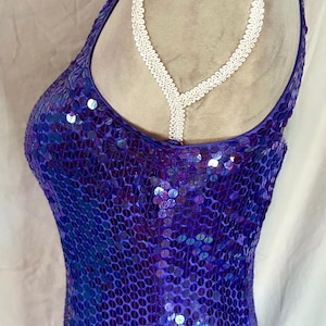 May include: A sleeveless, form-fitting dress covered in iridescent purple sequins. The dress has a halter neckline with a white, decorative trim. The fabric shimmers with shades of blue and purple, creating a dazzling effect.