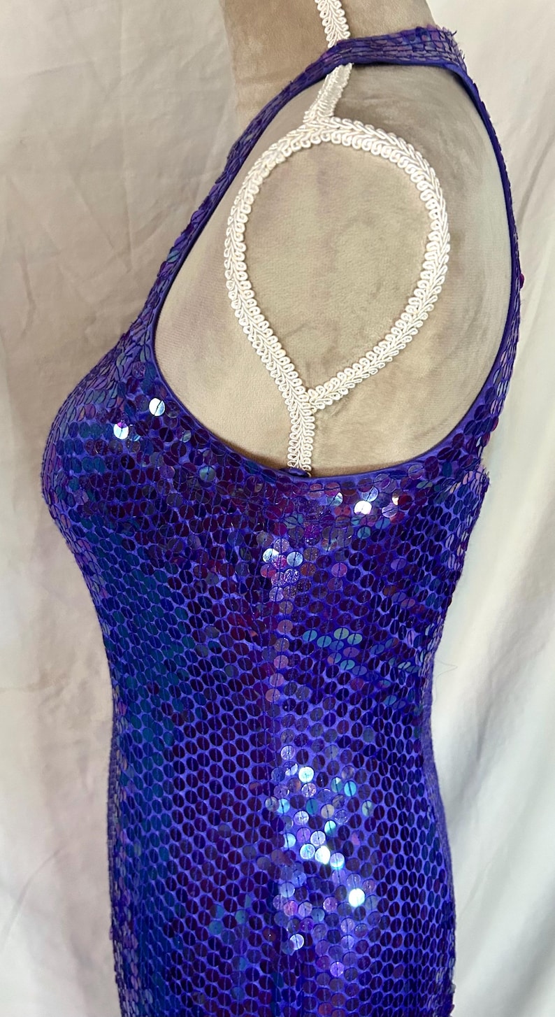 May include: A sleeveless, form-fitting dress covered in iridescent purple sequins. The dress has a halter neckline with a white, decorative trim. The fabric shimmers with shades of blue and purple, creating a dazzling effect.