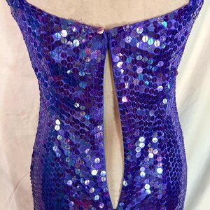 May include: A close-up of a purple sequined dress. The dress features a deep V-neckline and a fitted silhouette. The sequins are iridescent, reflecting shades of blue and purple. The dress appears to be made of a lightweight fabric.