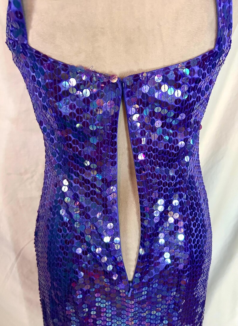 May include: A close-up of a purple sequined dress. The dress features a deep V-neckline and a fitted silhouette. The sequins are iridescent, reflecting shades of blue and purple. The dress appears to be made of a lightweight fabric.