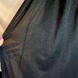 Puede incluir: Primer plano de una tela negra transparente con un sutil efecto brillante. La tela se sostiene, revelando su delicada textura y la forma en que la luz la atraviesa. El fondo es de color beige neutro.