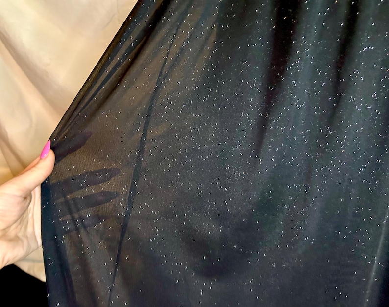 Puede incluir: Primer plano de una tela negra transparente con un sutil efecto brillante. La tela se sostiene, revelando su delicada textura y la forma en que la luz la atraviesa. El fondo es de color beige neutro.