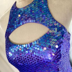 May include: A sleeveless, sequined garment in shades of purple and blue. The top features a keyhole cutout at the neckline. The sequins create a shimmering effect, reflecting light. The garment appears to be a dance costume or formal wear.