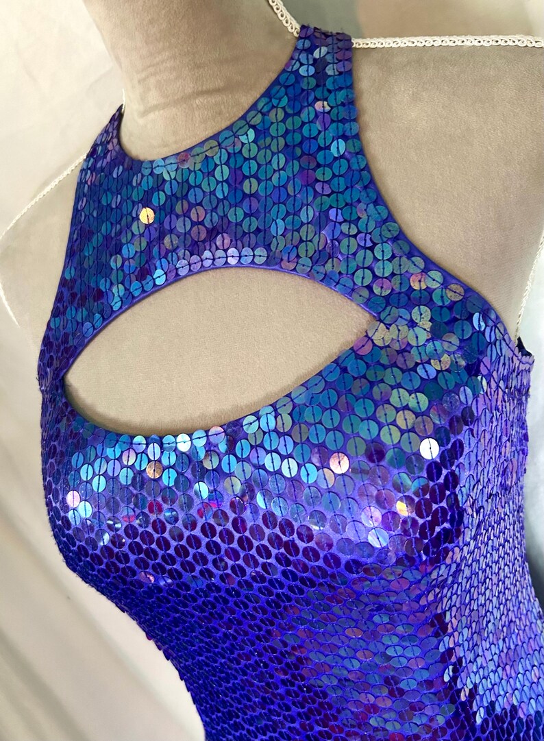 May include: A sleeveless, sequined garment in shades of purple and blue. The top features a keyhole cutout at the neckline. The sequins create a shimmering effect, reflecting light. The garment appears to be a dance costume or formal wear.
