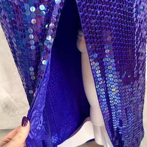 May include: A vibrant, floor-length gown covered in iridescent purple sequins. The dress features a high slit, revealing a deep blue lining. The fabric shimmers with a multitude of colors, creating a dazzling effect. The dress is displayed on a white mannequin.