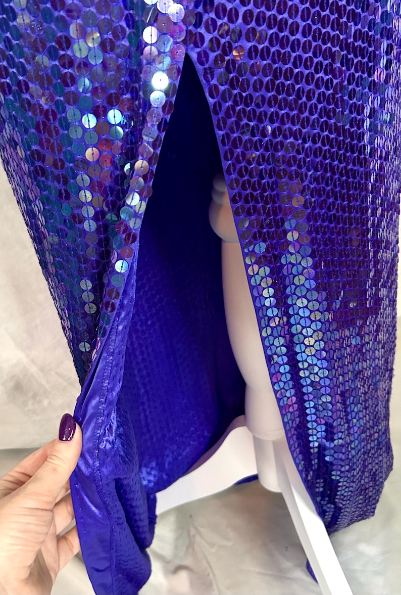 May include: A vibrant, floor-length gown covered in iridescent purple sequins. The dress features a high slit, revealing a deep blue lining. The fabric shimmers with a multitude of colors, creating a dazzling effect. The dress is displayed on a white mannequin.