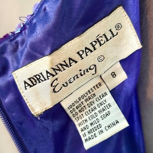 May include: Close-up of a label from an Adrianna Papell Evening dress. The label is white with black text, including the brand name, care instructions, and size 8. The dress is a vibrant blue with sequin details.