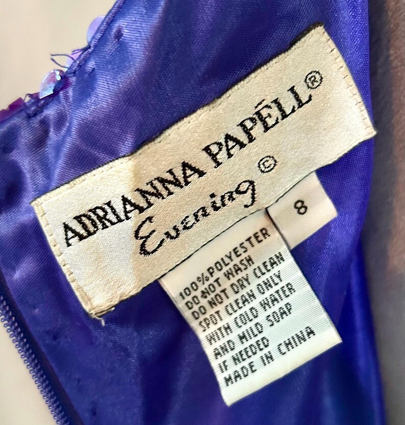 May include: Close-up of a label from an Adrianna Papell Evening dress. The label is white with black text, including the brand name, care instructions, and size 8. The dress is a vibrant blue with sequin details.