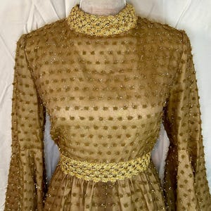 May include: A vintage gold-colored dress with long sleeves and a high, embellished collar. The dress features a textured pattern and a decorative gold belt at the waist. The sleeves flare out from the elbow.