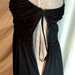 Puede incluir: Vestido de noche negro con una superposici&oacute;n transparente y brillante. El vestido presenta un corpi&ntilde;o fruncido, tirantes finos y una cremallera en la espalda. Un ribete de encaje blanco es visible en la parte posterior. El vestido se exhibe en un maniqu&iacute;.