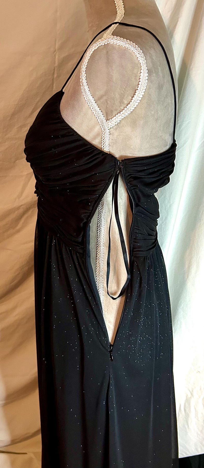 Puede incluir: Vestido de noche negro con una superposici&oacute;n transparente y brillante. El vestido presenta un corpi&ntilde;o fruncido, tirantes finos y una cremallera en la espalda. Un ribete de encaje blanco es visible en la parte posterior. El vestido se exhibe en un maniqu&iacute;.