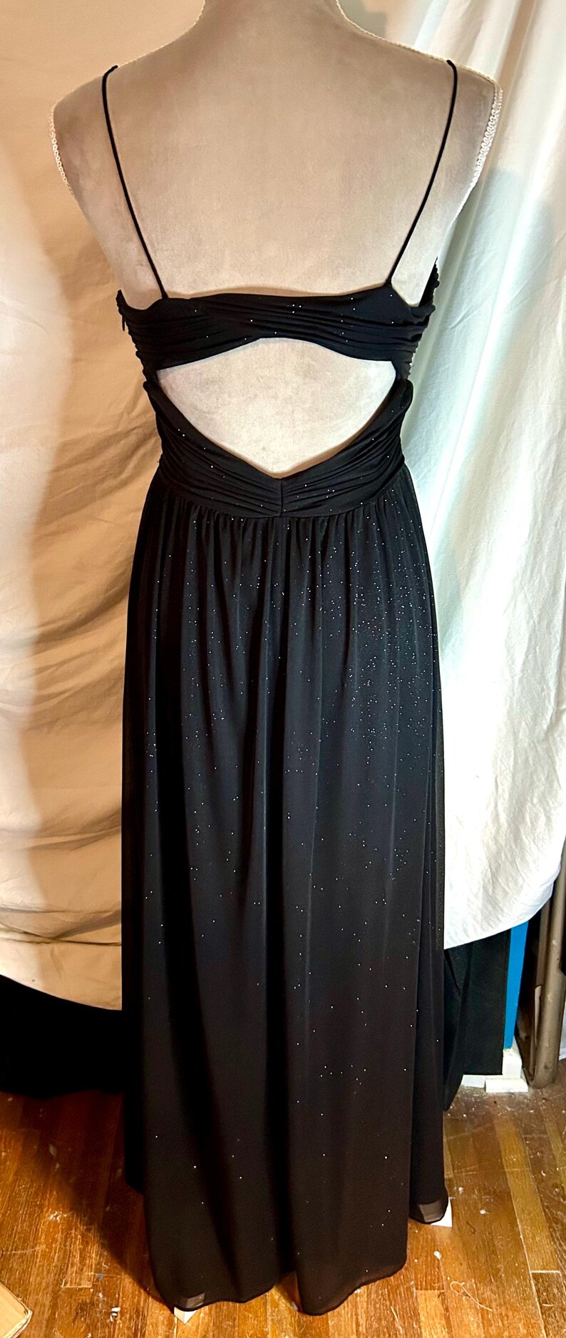 Puede incluir: Un vestido de noche negro con una falda fluida y tirantes finos. El vestido presenta una espalda descubierta y est&aacute; adornado con peque&ntilde;os detalles brillantes en toda la tela. El vestido se exhibe en un maniqu&iacute;.