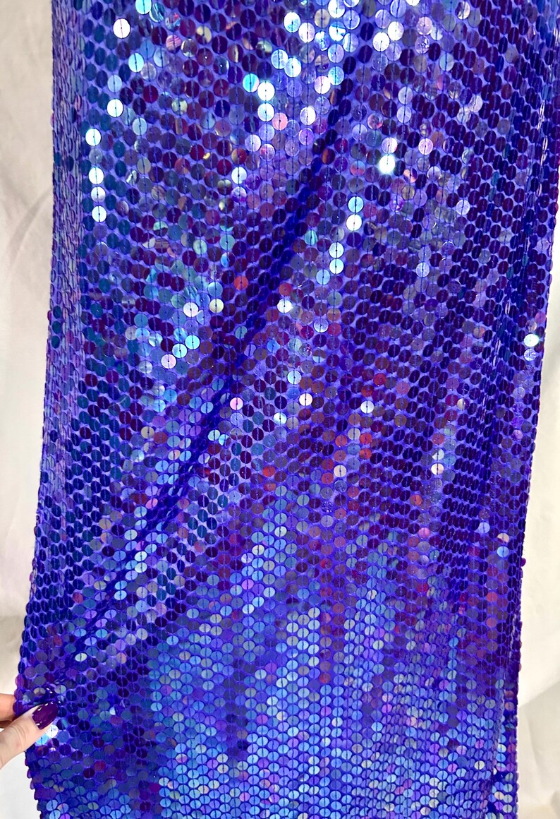 May include: A close-up of a fabric covered in iridescent blue and purple sequins. The sequins are round and densely packed, creating a shimmering effect. The fabric is likely used for apparel or decorative purposes.