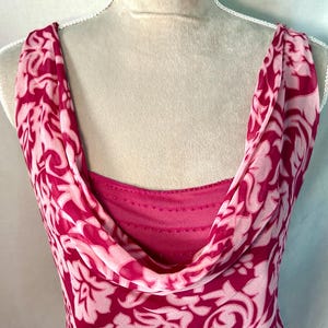 May include: A sleeveless dress featuring a cowl neckline and a layered design. The outer layer is a sheer fabric with a bold magenta and light pink floral pattern, while the inner layer is a solid magenta fabric with horizontal stitching details.