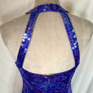 May include: A close-up of a blue sequined halter dress. The dress features a high collar and straps that extend to the back. The sequins are a mix of blue and iridescent colors, creating a shimmering effect. The dress is on a mannequin.