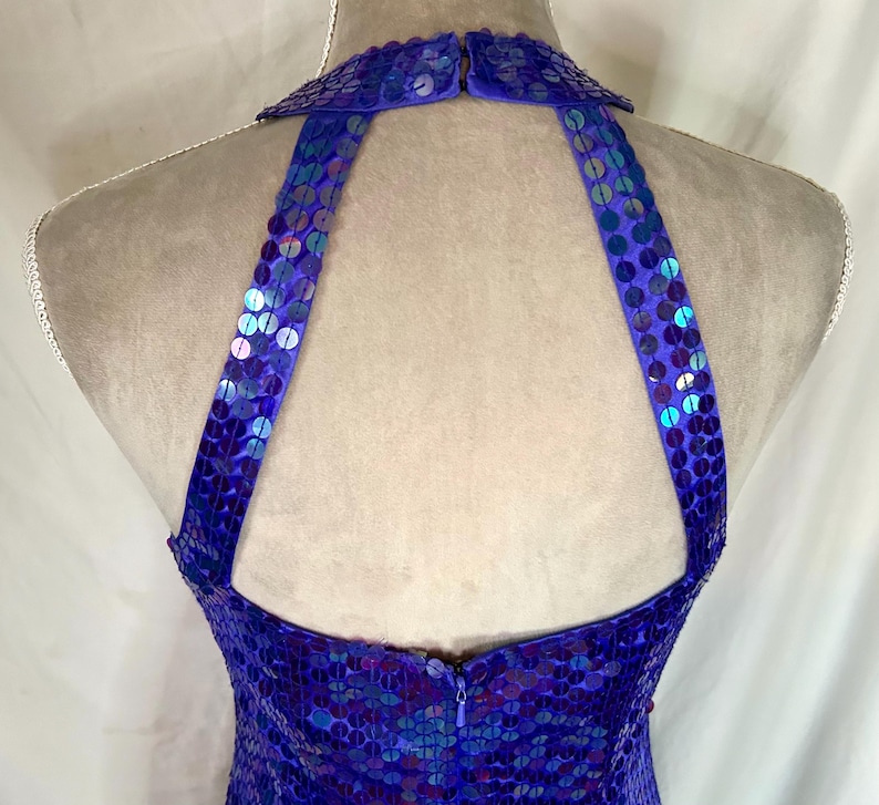 May include: A close-up of a blue sequined halter dress. The dress features a high collar and straps that extend to the back. The sequins are a mix of blue and iridescent colors, creating a shimmering effect. The dress is on a mannequin.