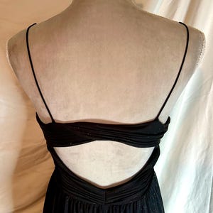 Puede incluir: Vestido de noche negro con tirantes finos y espalda baja. El corpi&ntilde;o presenta un dise&ntilde;o fruncido con sutiles detalles brillantes. El vestido se exhibe en un maniqu&iacute;, mostrando su elegante silueta y tejido fluido.