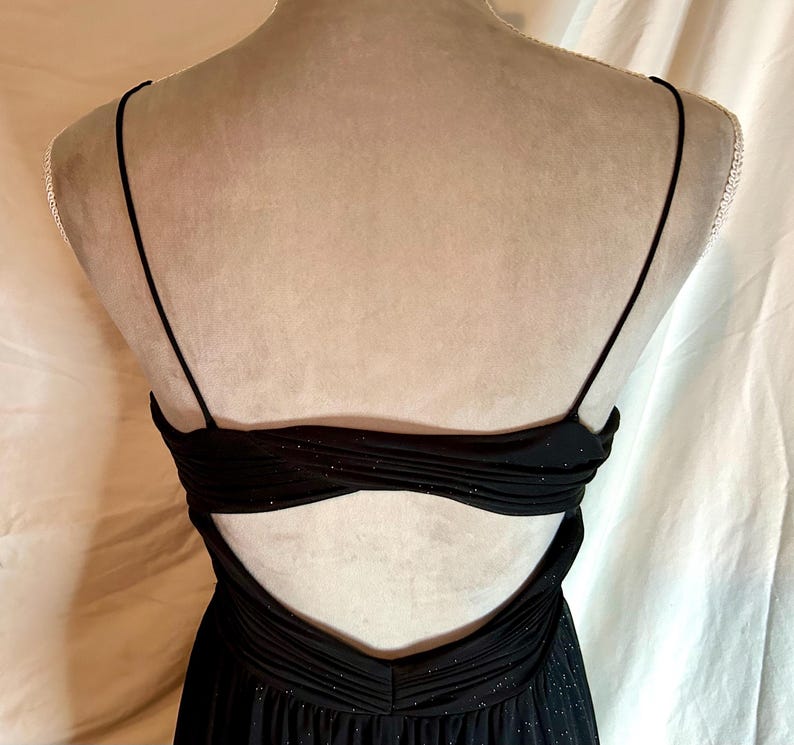Puede incluir: Vestido de noche negro con tirantes finos y espalda baja. El corpi&ntilde;o presenta un dise&ntilde;o fruncido con sutiles detalles brillantes. El vestido se exhibe en un maniqu&iacute;, mostrando su elegante silueta y tejido fluido.