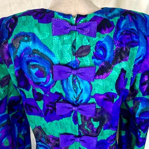 May include: A vintage dress back with a vibrant floral pattern in shades of teal, blue, and purple. The dress features a series of purple bow accents down the back. The fabric has a textured appearance.