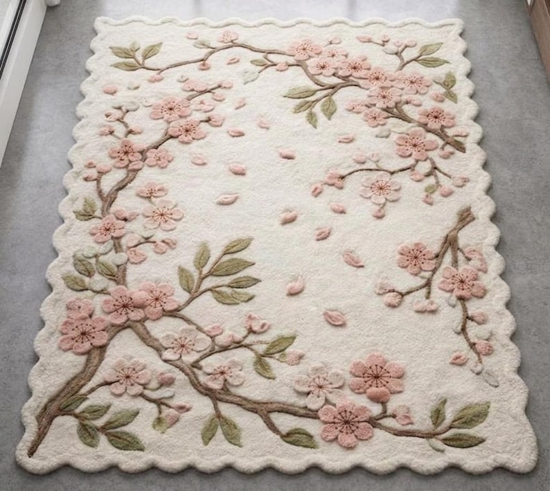 May include: Cream-colored rectangular rug with a scalloped edge, featuring a 3D floral design. Pink cherry blossoms and falling petals are arranged on brown branches with green leaves, creating a nature-inspired home decor piece.
