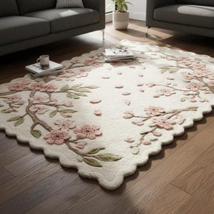 May include: Off-white rug with a scalloped edge, featuring a floral design of pink blossoms and green leaves on brown branches. The rug is placed on a hardwood floor, creating a soft and inviting aesthetic.