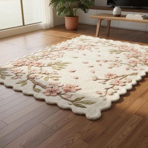 May include: Cream-colored rectangular rug with a scalloped edge, featuring a floral design of pink blossoms and green leaves on brown branches. The rug is placed on a hardwood floor.