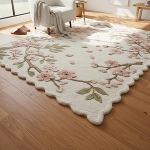 May include: A cream-colored rug with a floral design featuring pink blossoms and green leaves on brown branches. The rug has a scalloped edge and is placed on a hardwood floor. The rug is a decorative home decor item.