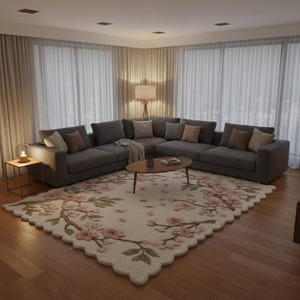 May include: A modern living room featuring a large gray sectional sofa with several throw pillows. A round wooden coffee table sits on a floral rug with pink blossoms. The room has large windows with sheer curtains and a side table with a lamp.