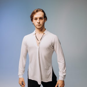 Op de afbeelding: Een lichtgrijs shirt met lange mouwen, een opstaande kraag en een splitzoom, gecombineerd met een zwarte broek. De bovenste knoop van het shirt is open, waardoor een ketting zichtbaar is. De achtergrond is een blauwe gradiënt.