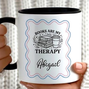 May include: White ceramic mug with a black interior and handle. The mug features a design with the text "BOOKS ARE MY THERAPY" and the name "Abigail". The design includes illustrations of books and a coffee cup.